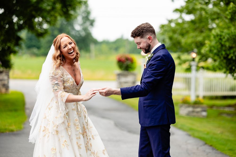 Bride in floral gown laughing joyfully as groom in navy suit holds her hand outdoors at wedding ceremony.