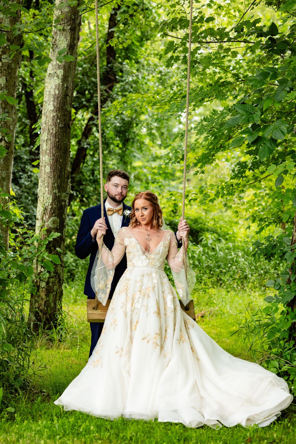 Elegant bride in floral gown sitting on a wooden swing as groom in navy suit stands behind her.
