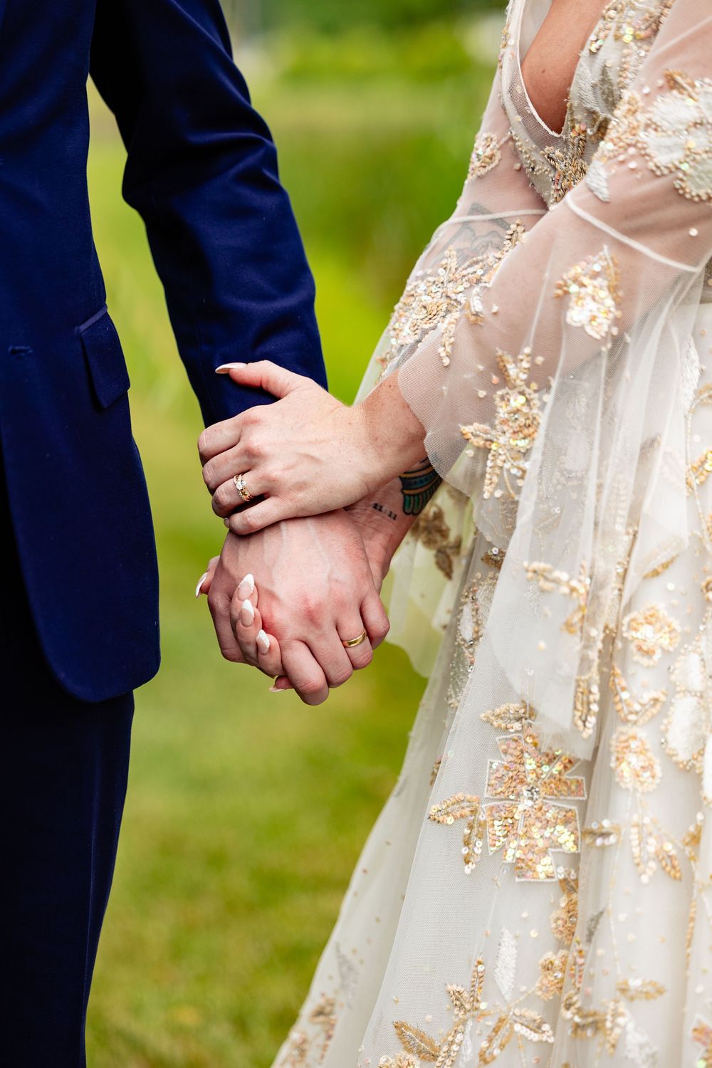 Bride and groom holding hands, showcasing wedding rings, gold embroidered bridal gown, and navy blue suit at outdoor ceremony.