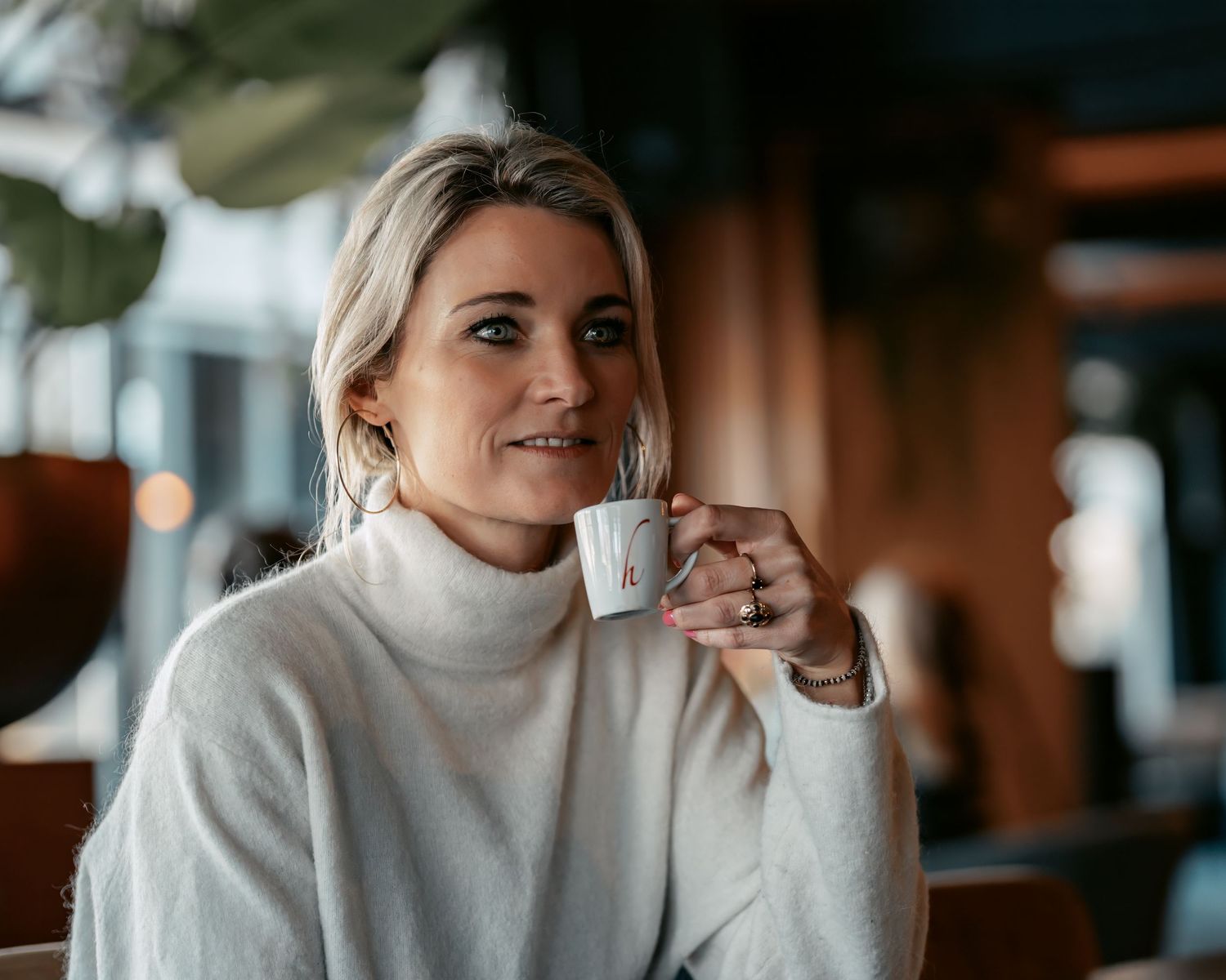 Person in cozy white turtleneck sweater holding coffee cup in warm indoor setting.