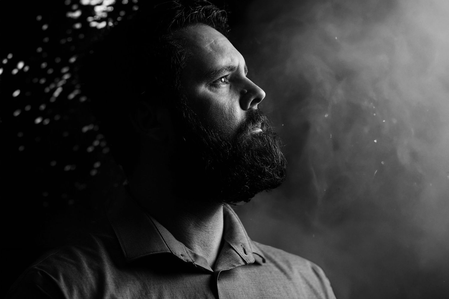 Dramatic black and white profile portrait of a bearded man gazing thoughtfully into misty darkness.