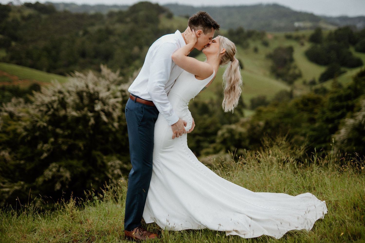 A couple shares a romantic kiss on a hilltop overlooking rolling green countryside in their wedding attire.