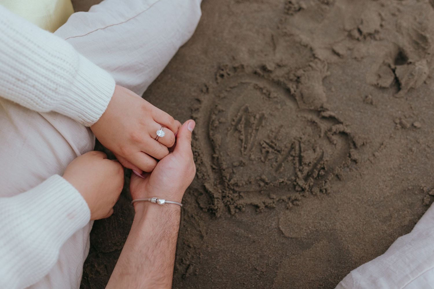close up of a couple holding hands on a beach with letters "N+N" within a heart written in the sand