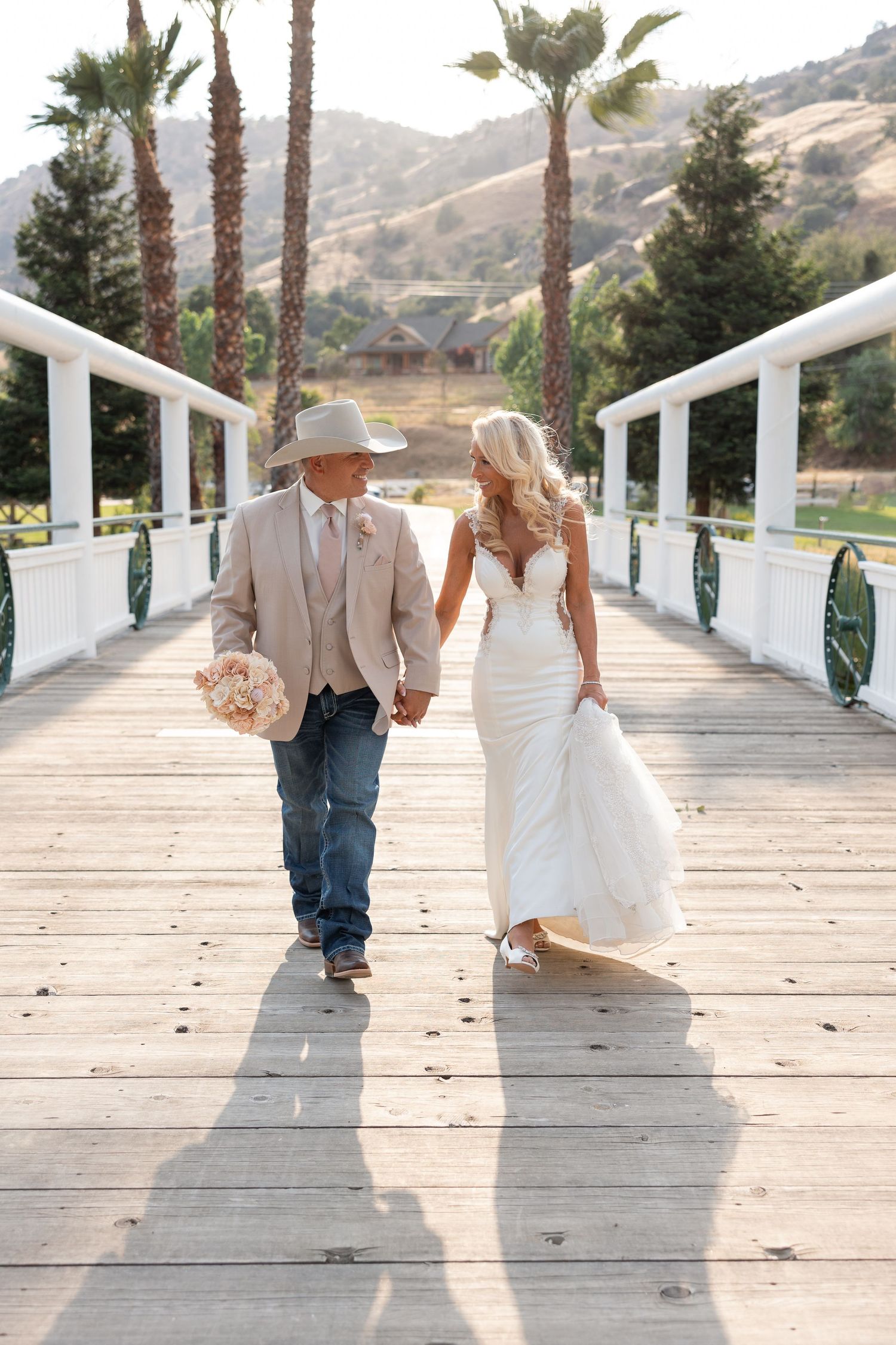 Couple in wedding attire walking hand and hands across wooden bridge at springville ranch with palm trees and hills in the background.