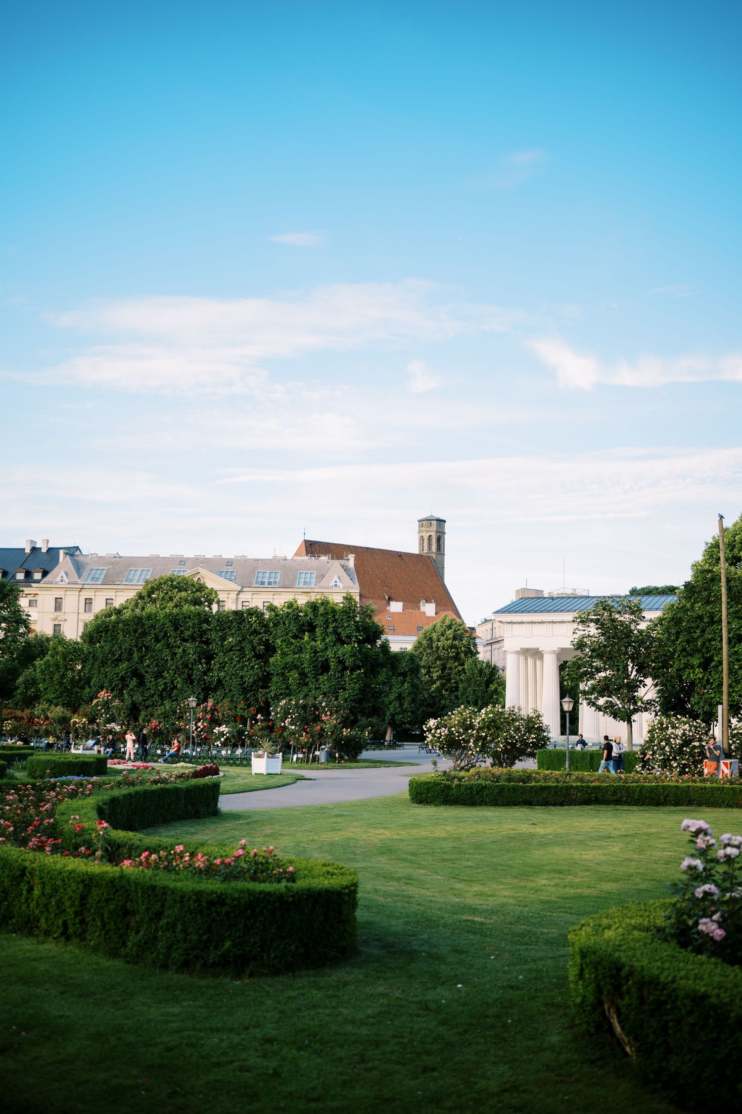 A manicured garden with green hedges and lawns in front of classical buildings under a bright blue sky.