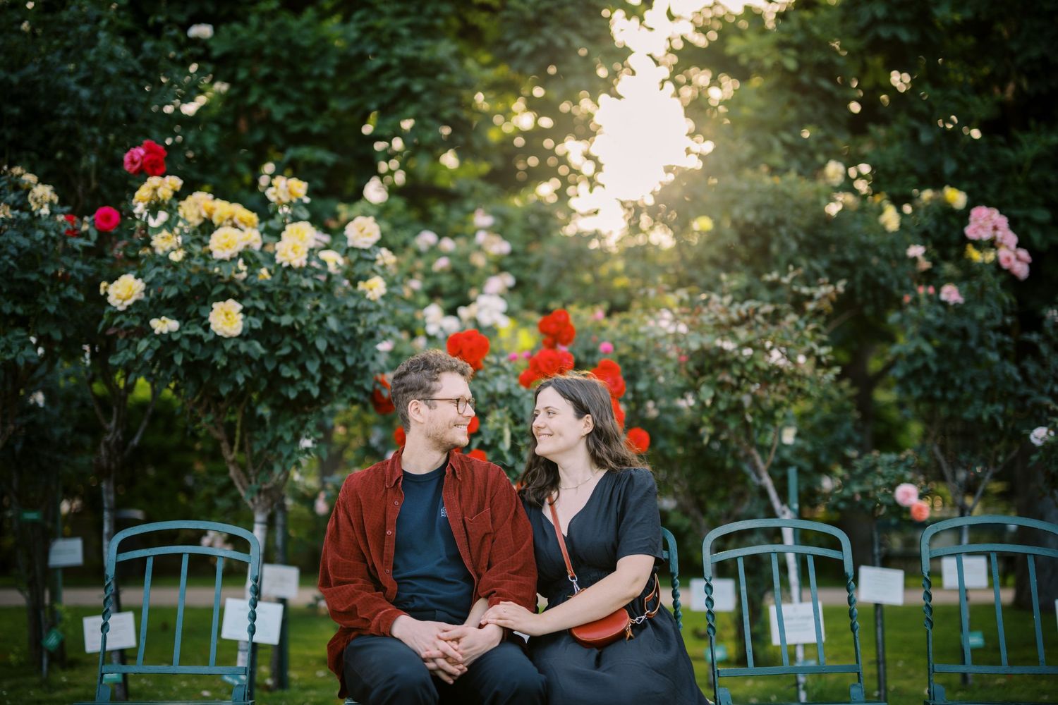 A couple sits on a bench in a romantic garden filled with colorful roses and blue chairs at sunset.
