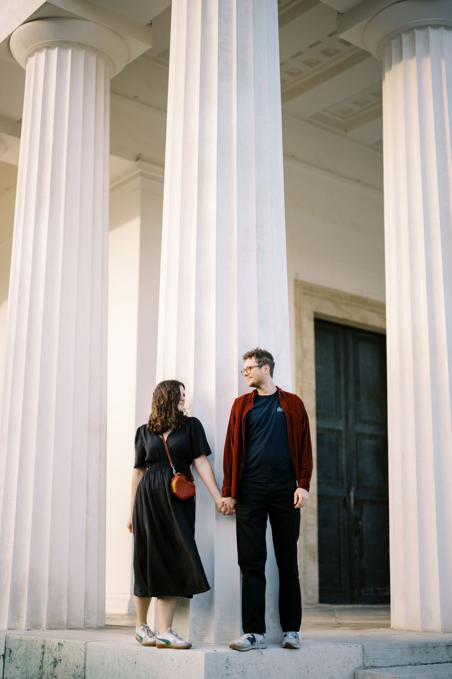 A couple stands holding hands between tall white columns on the steps of a classical building.