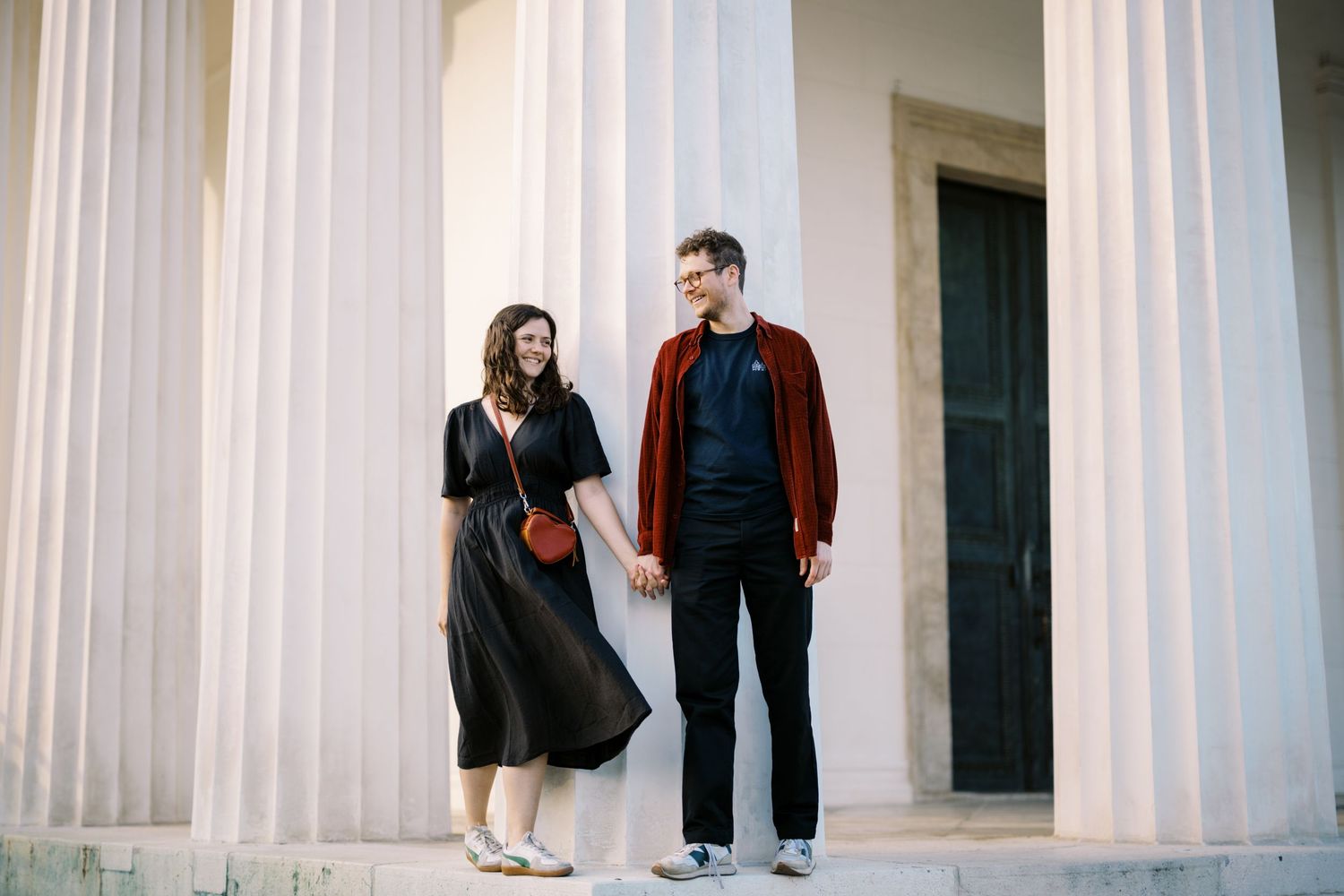 Two people stand between white classical columns while sharing a moment on a bright day.