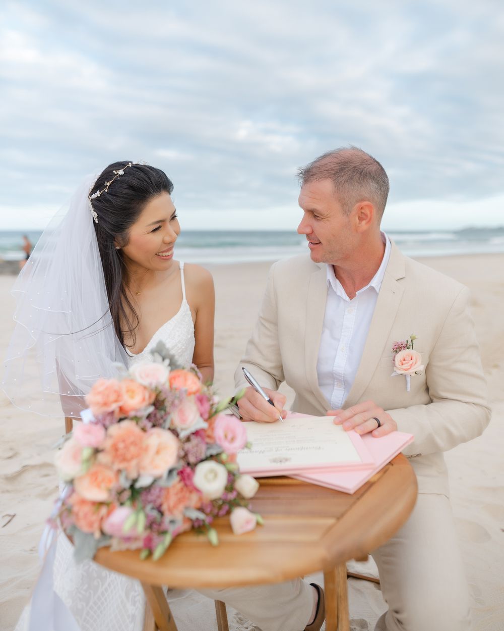 Beach wedding ceremony with couple signing marriage documents next to coral flower bouquet.