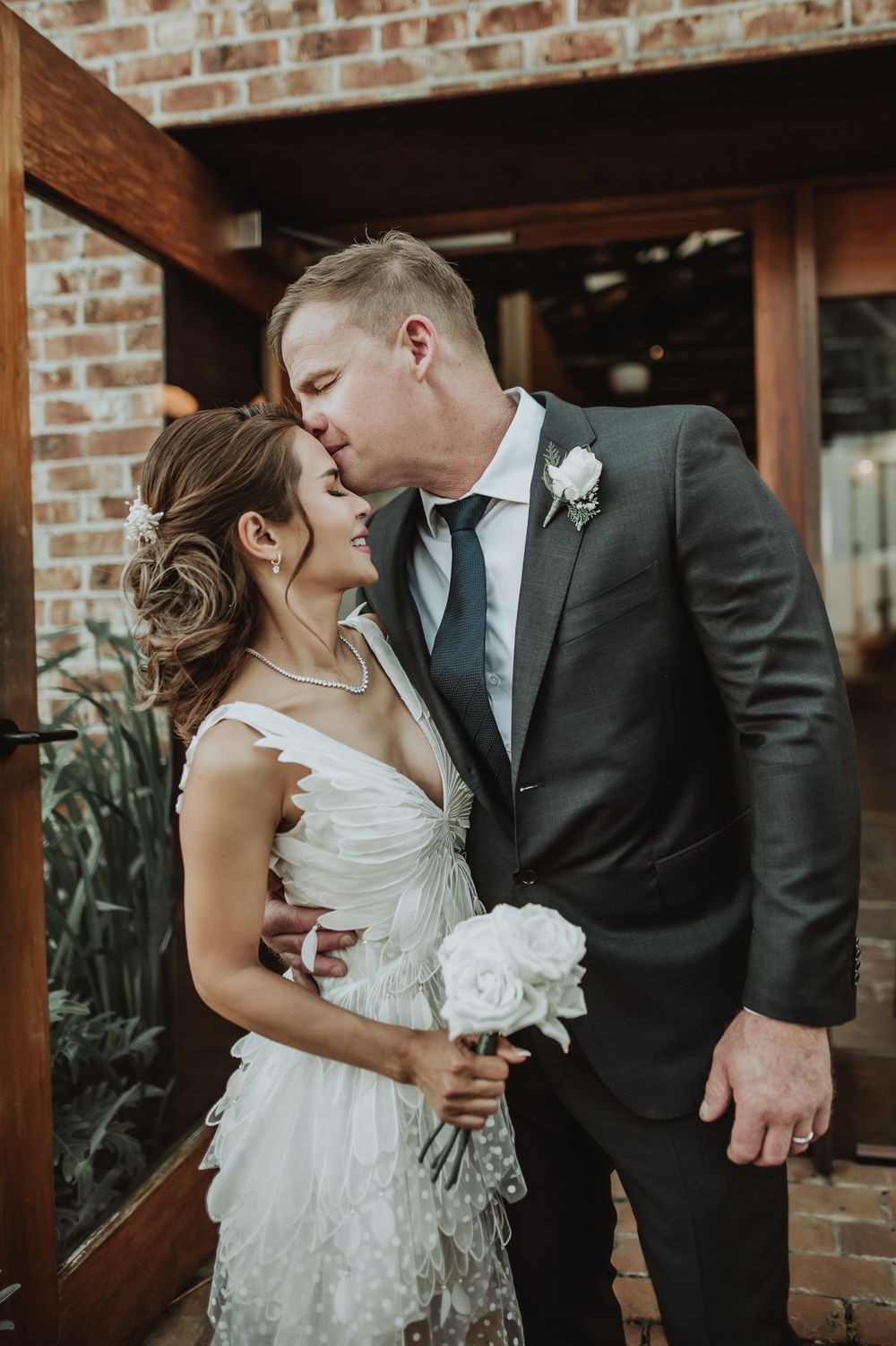 A romantic wedding moment captured against a rustic brick wall background with white floral bouquet details.