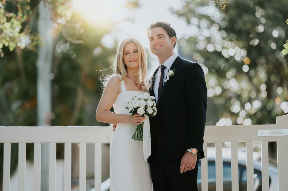 Newlyweds stand together on white porch holding bouquet in outdoor garden setting.