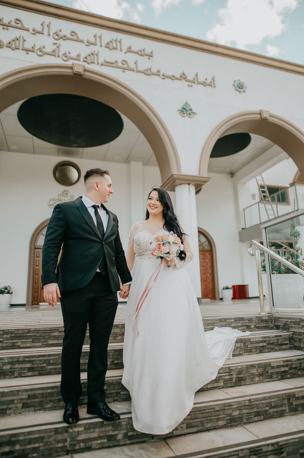 Newlyweds pose on steps of mosque with Arabic script and arched architecture.