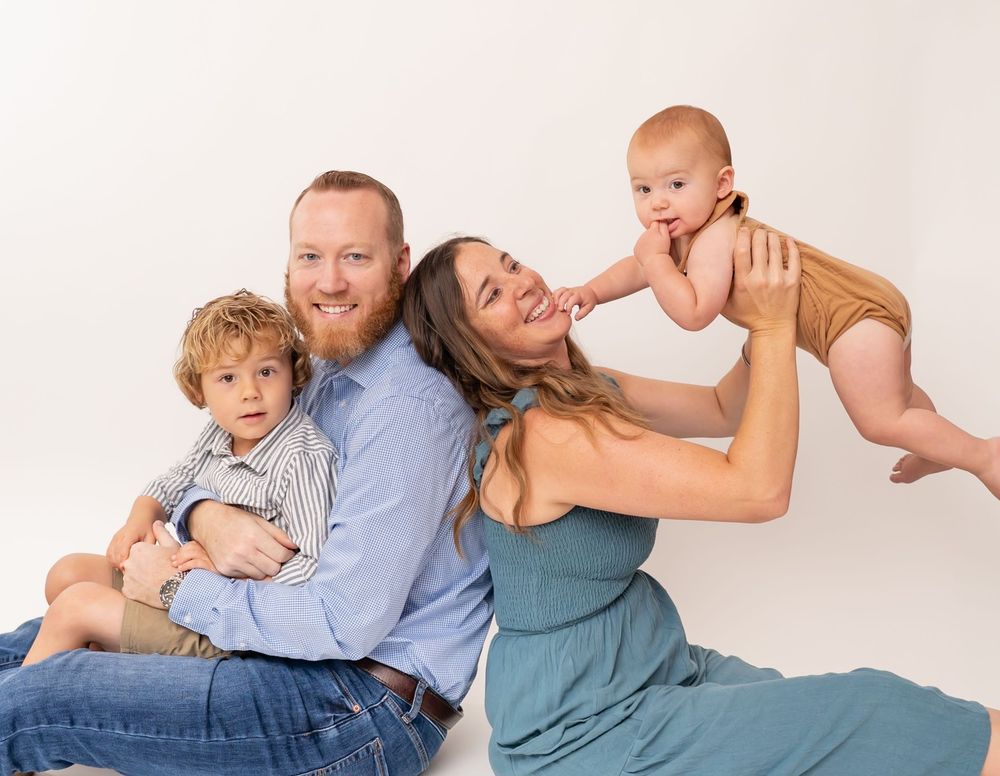 Happy family of four posing in studio, parents sitting with toddler son and mother holding smiling baby in tan outfit.
