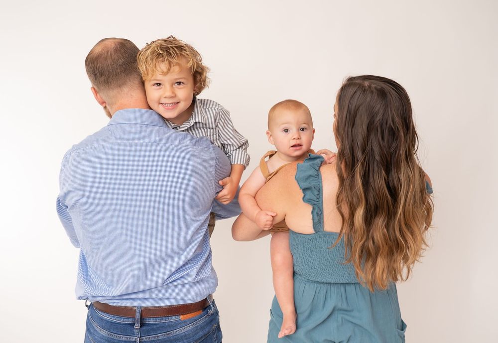 A family of four poses against a white backdrop, parents holding a toddler boy and baby in a warm studio portrait.