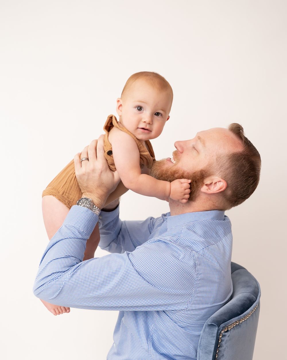 A father lifts his smiling baby in a bright studio, both wearing blue and tan outfits.