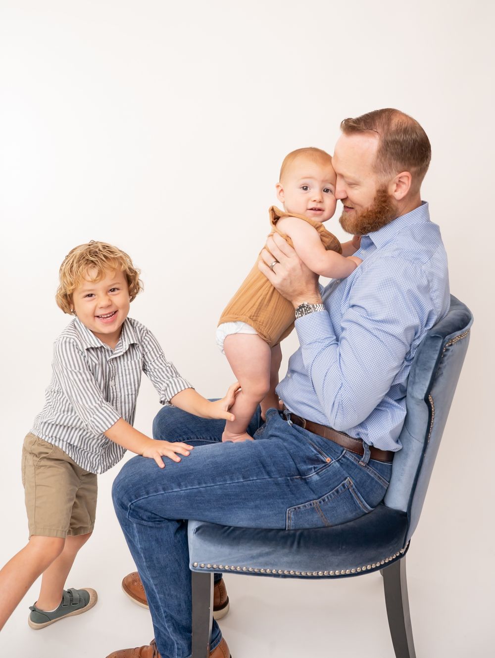 A red-bearded father sits on a blue chair holding an infant while his young son laughs beside him.