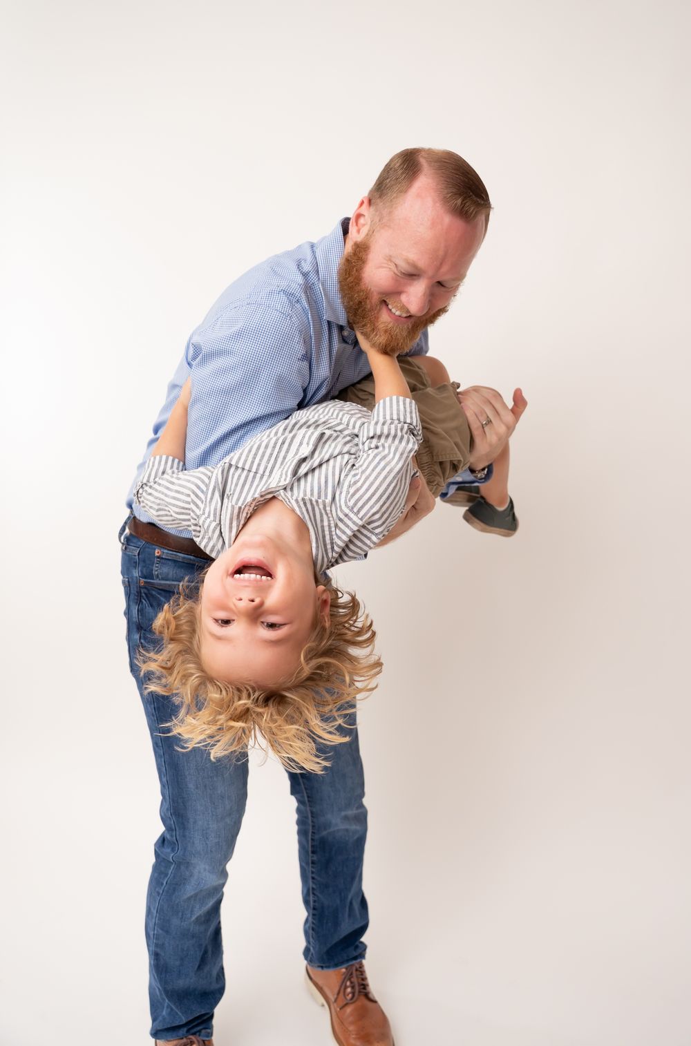 A bearded father playfully holds his laughing blond child upside down against a white studio background.