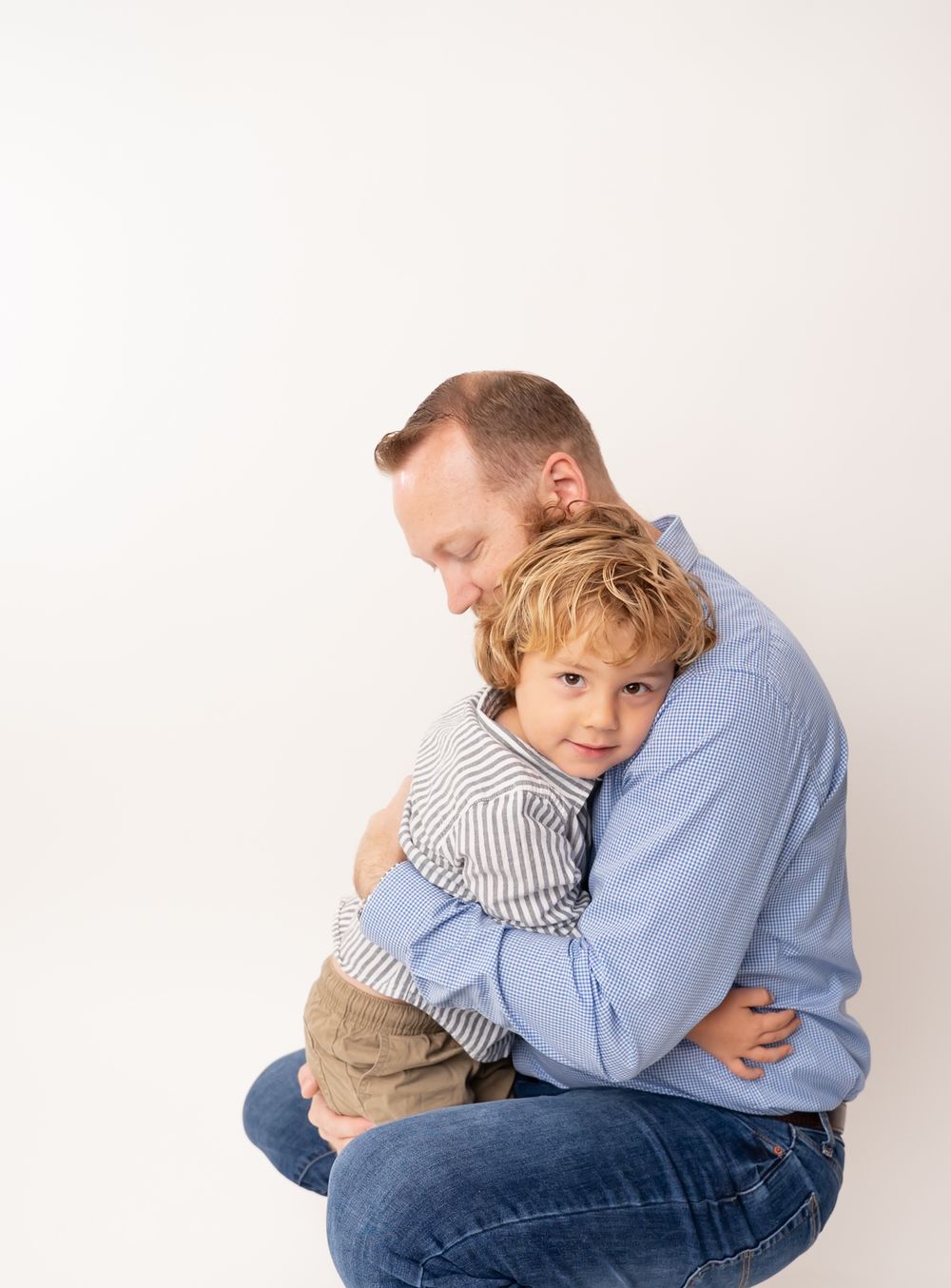 A father hugs his young blond son tenderly in a clean white studio setting.