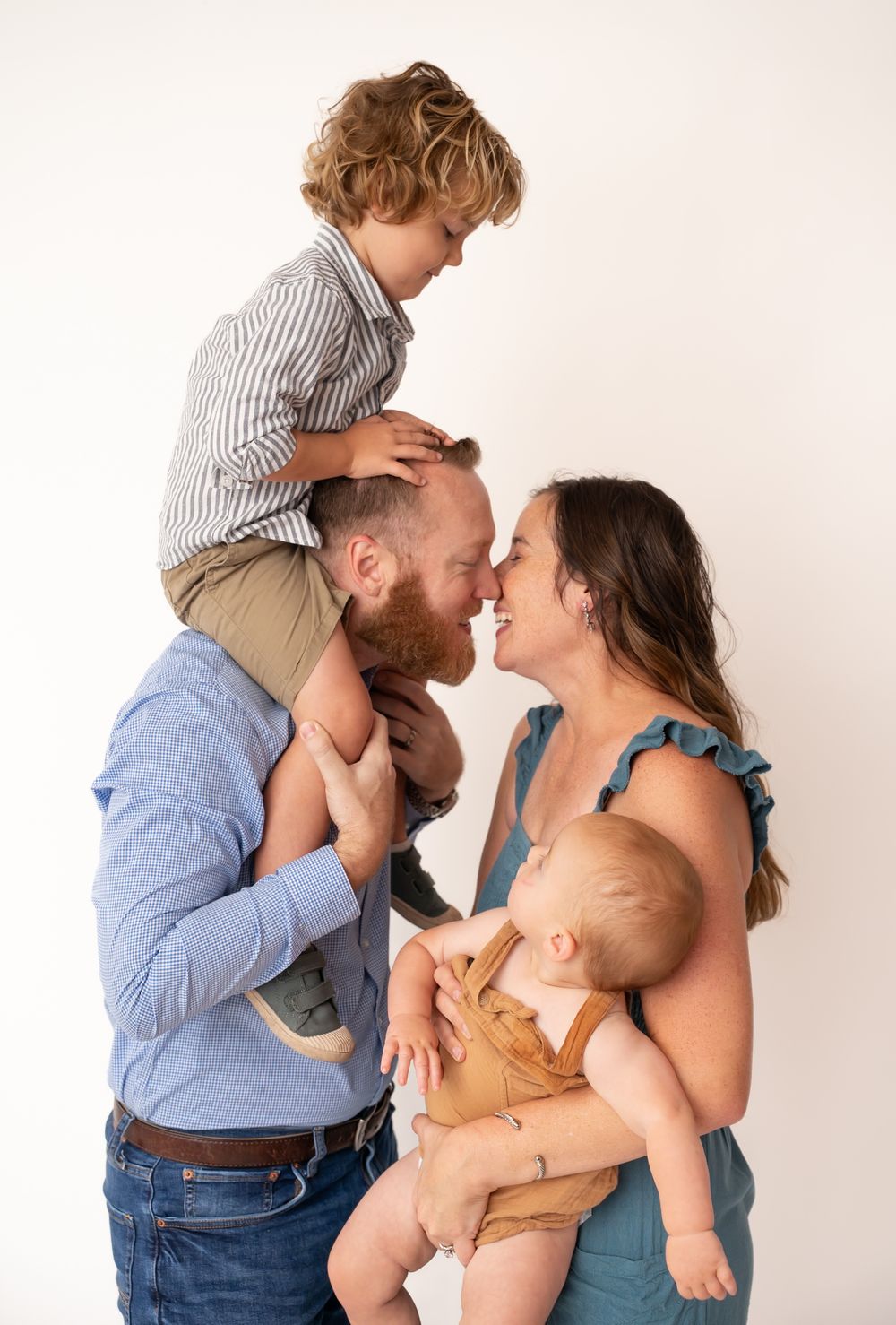 A happy family of four posing together, with dad holding toddler on shoulders and mom cradling baby in a bright studio.
