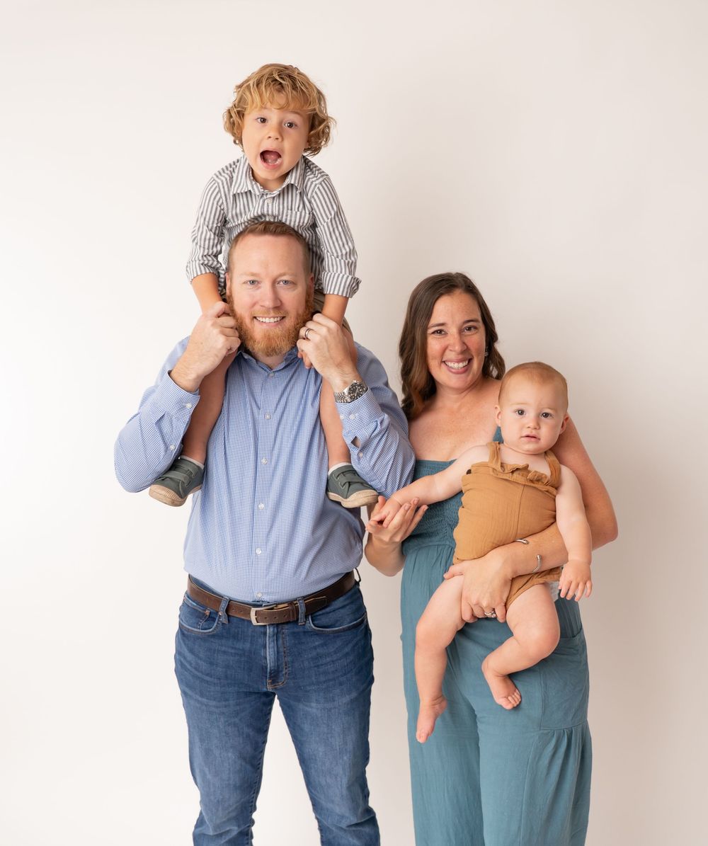 Happy family portrait with dad, toddler on shoulders, mom holding baby, smiling against white background.