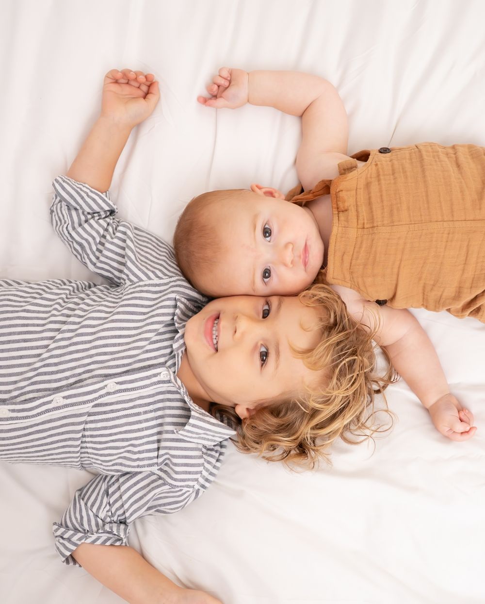 Two young siblings, a toddler and baby, lying together on white bedding, smiling cutely at the camera.