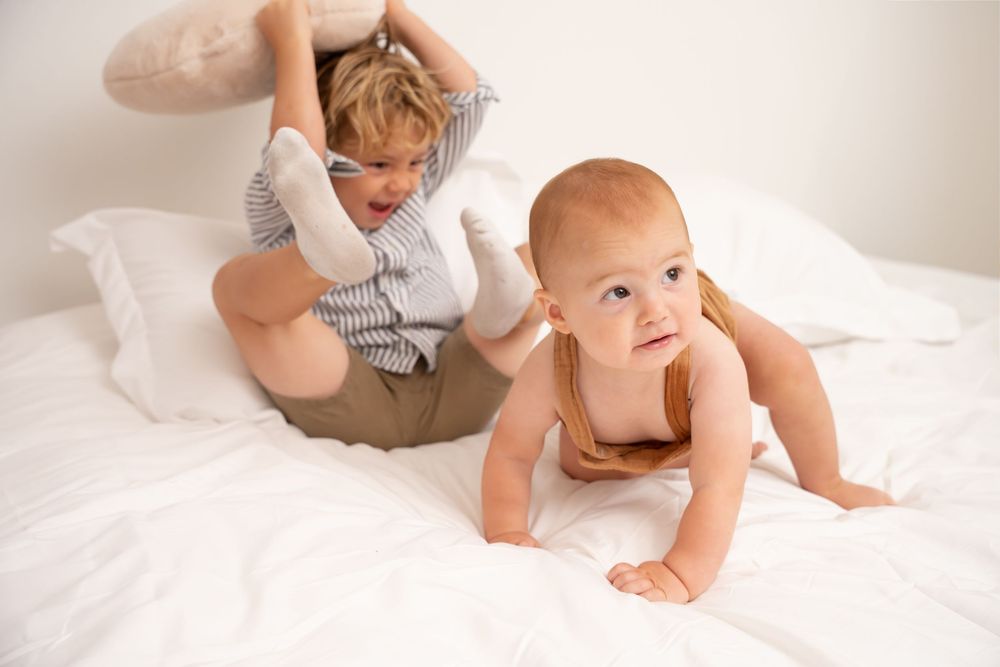 A curious baby crawls on a white bed while an older sibling plays nearby in a candid family photo.