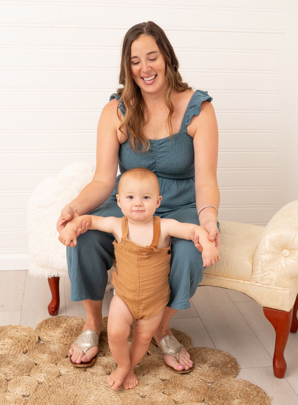 A smiling woman in a teal jumpsuit sits on a bench helping a baby in tan overalls stand on a woven rug.