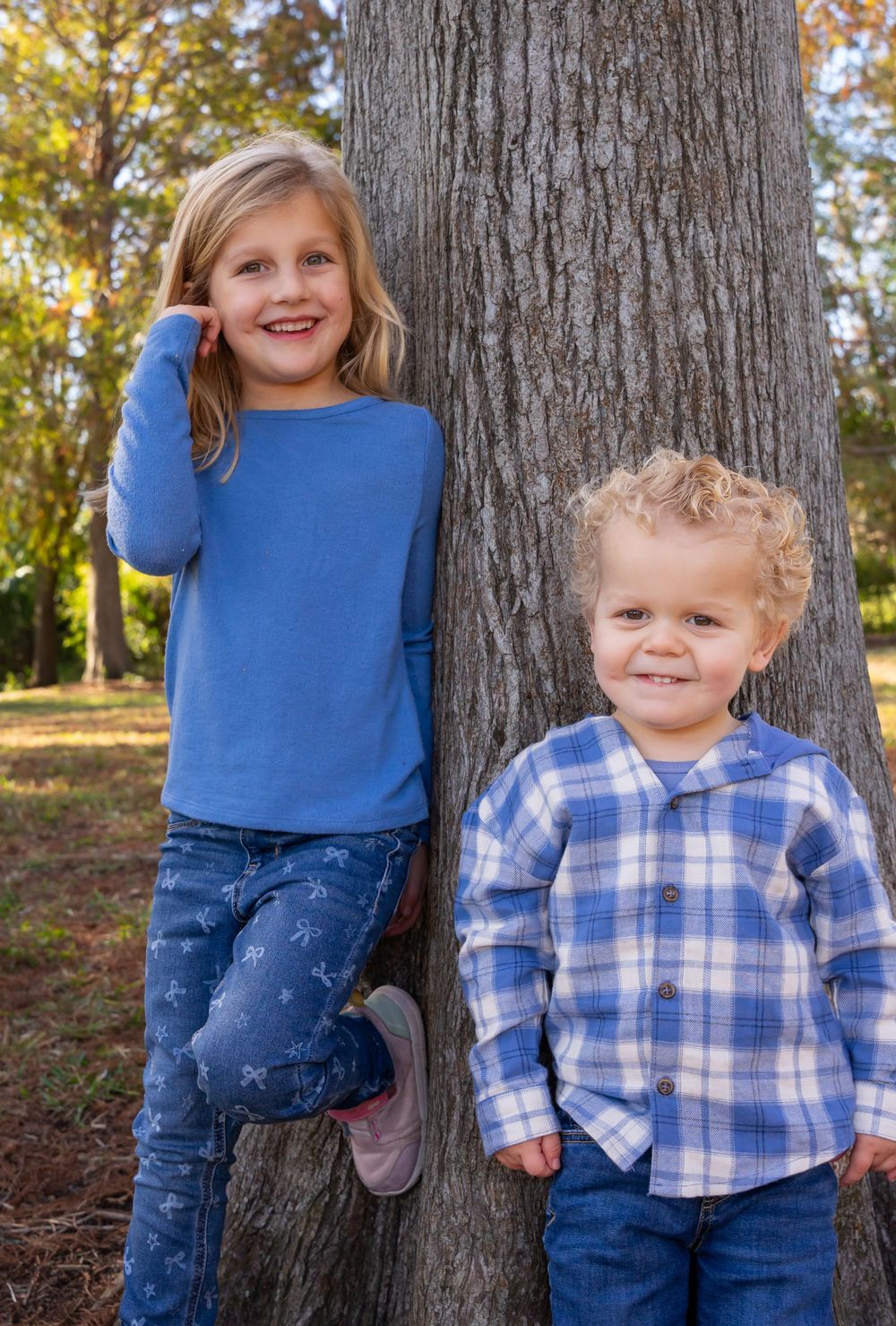 Two children posing against a large tree trunk wearing coordinating blue outfits.