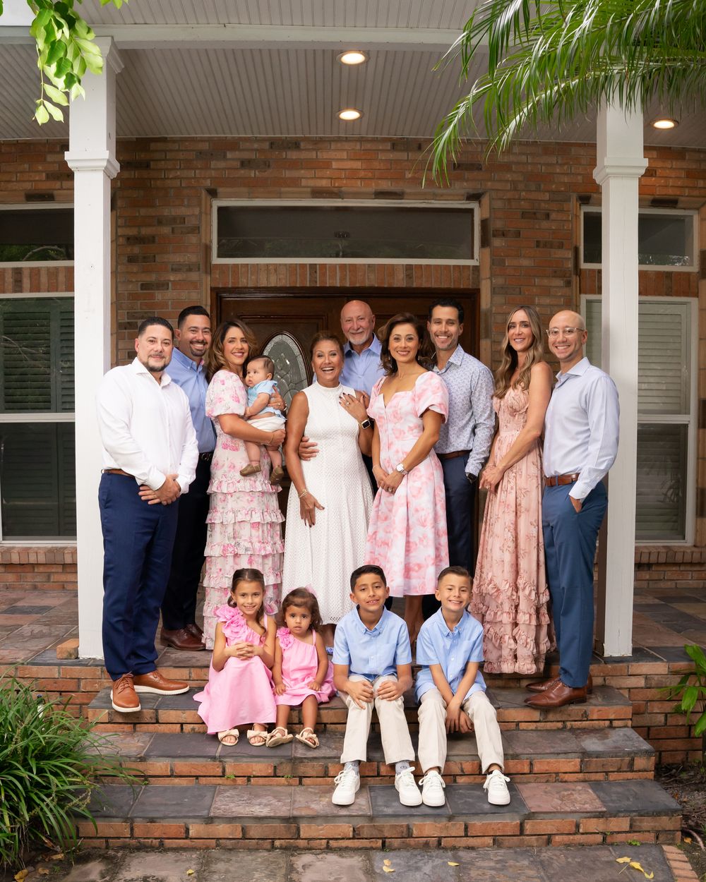 A large multigenerational family smiles together on the brick steps of a home, dressed in elegant pastel attire.