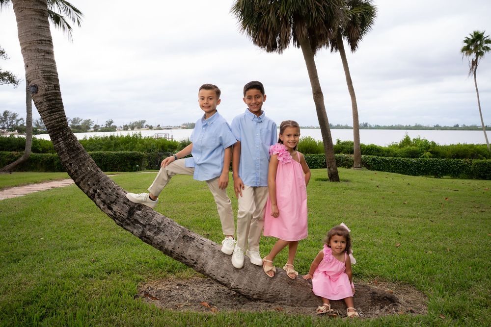 Four children posing on a leaning palm tree in a tropical park setting near the water.