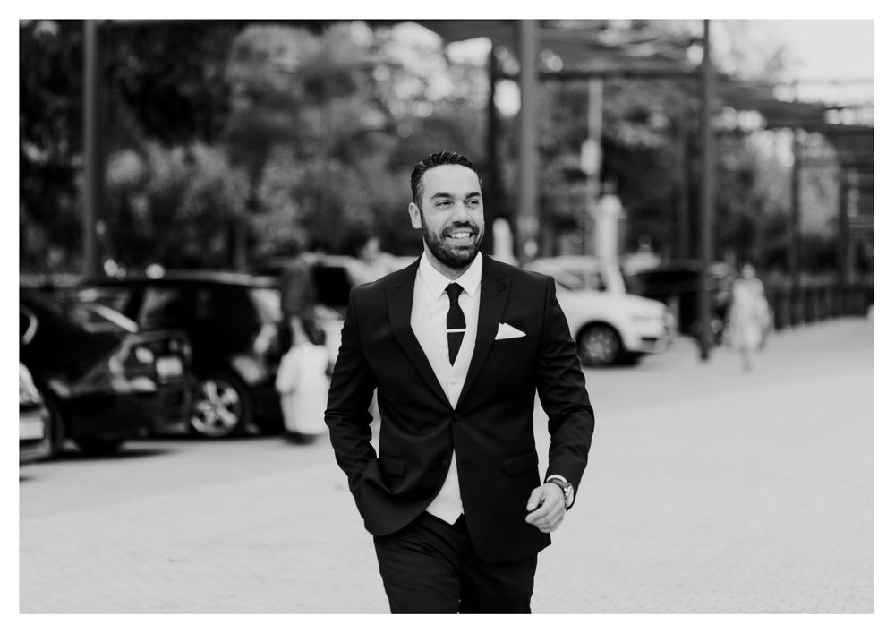 Groom in classic black suit poses confidently in black and white street portrait.