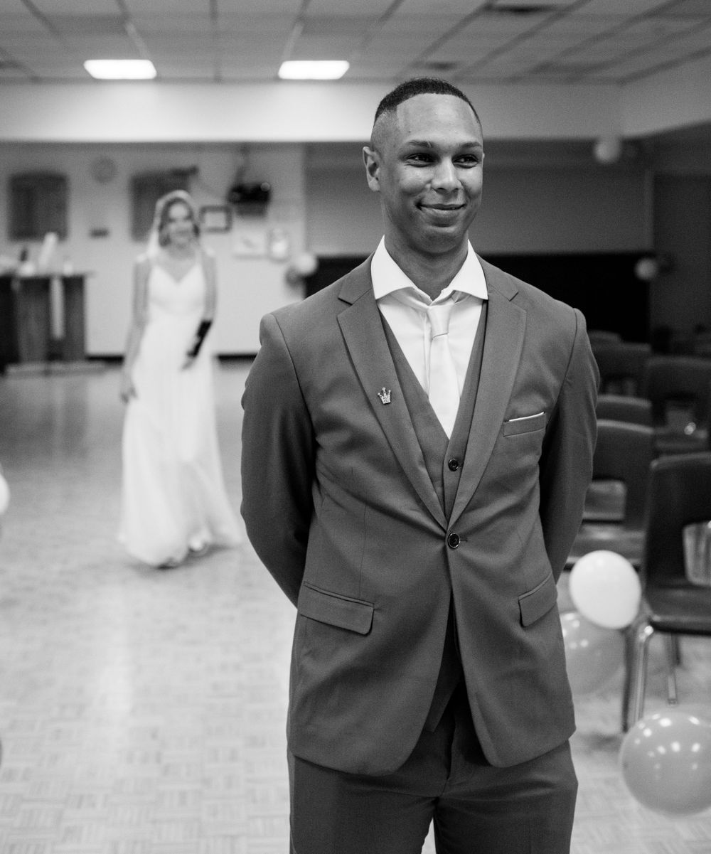 A black and white portrait of a groom in a formal suit with a figure in white visible in the background.