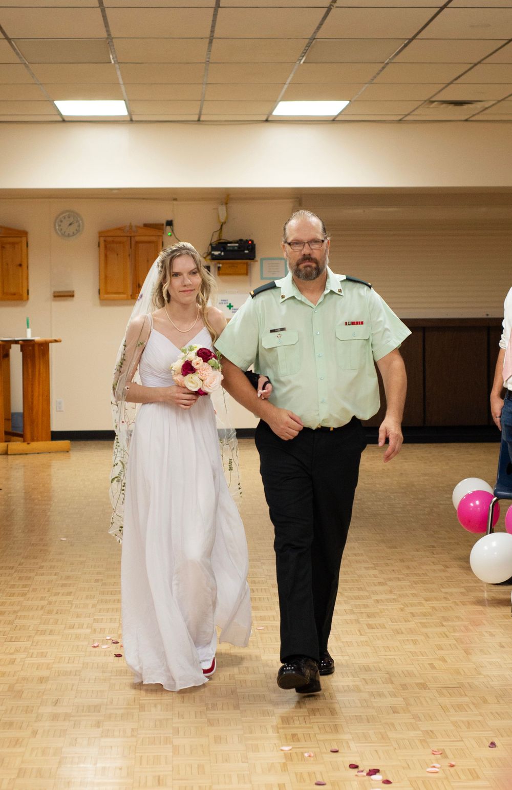 A bride in a white dress walking down the aisle escorted by a person in a green service uniform at an indoor venue.