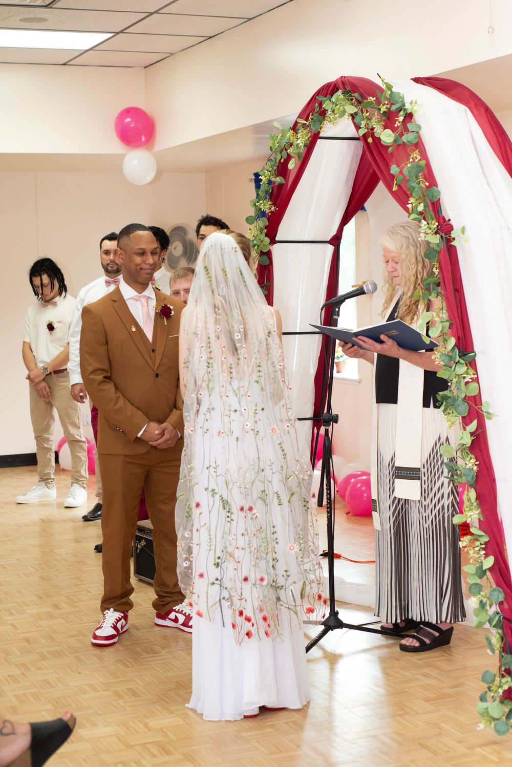 Wedding ceremony in progress under decorated arch with pink balloons and floral details.