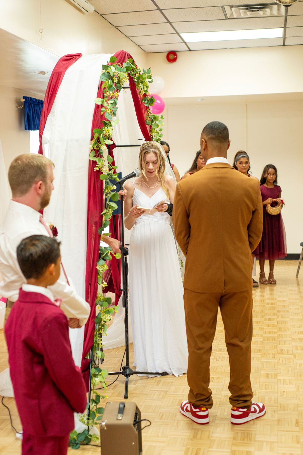 A wedding ceremony takes place under a decorated floral arch in an indoor venue with red and white decor.