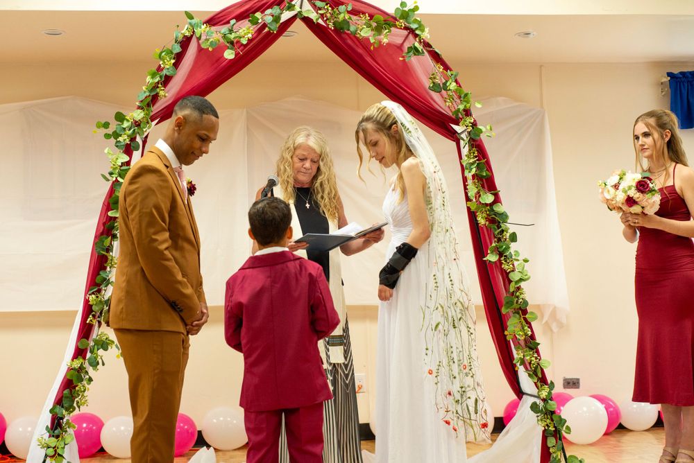 A wedding ceremony takes place under a red floral arch with celebrants gathered for the intimate indoor occasion.