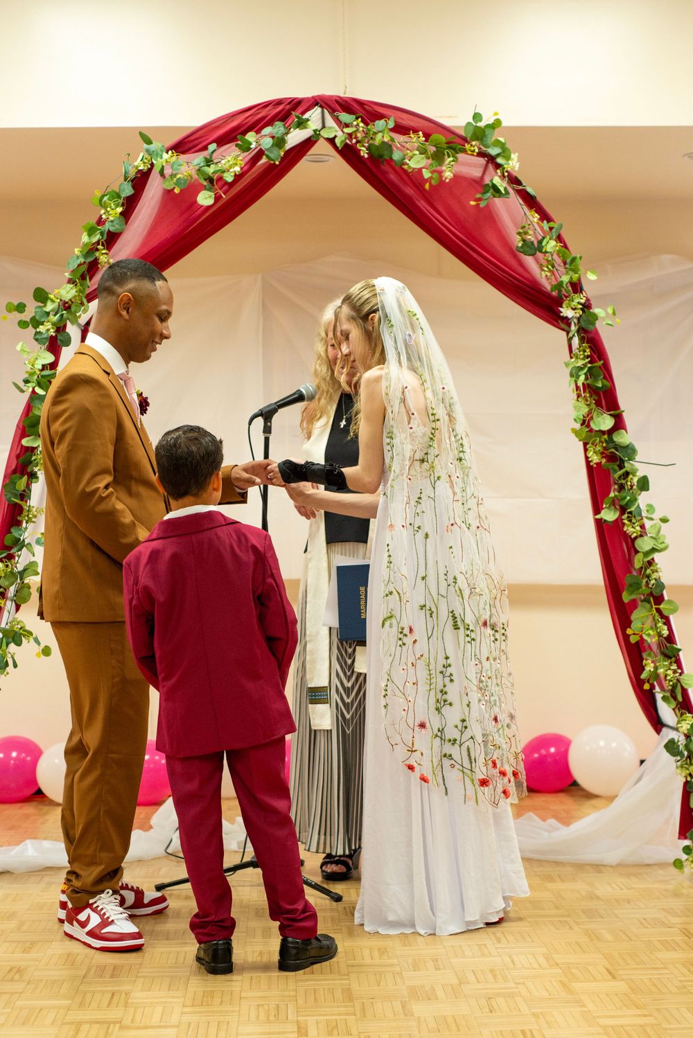 Wedding ceremony under red arch with greenery and pink balloons in indoor venue.