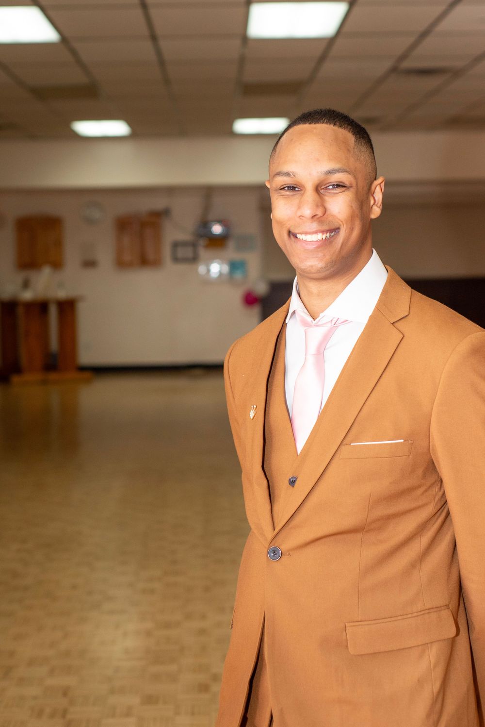 A confident gentleman in a tan suit and pink dress shirt smiles in an indoor setting.