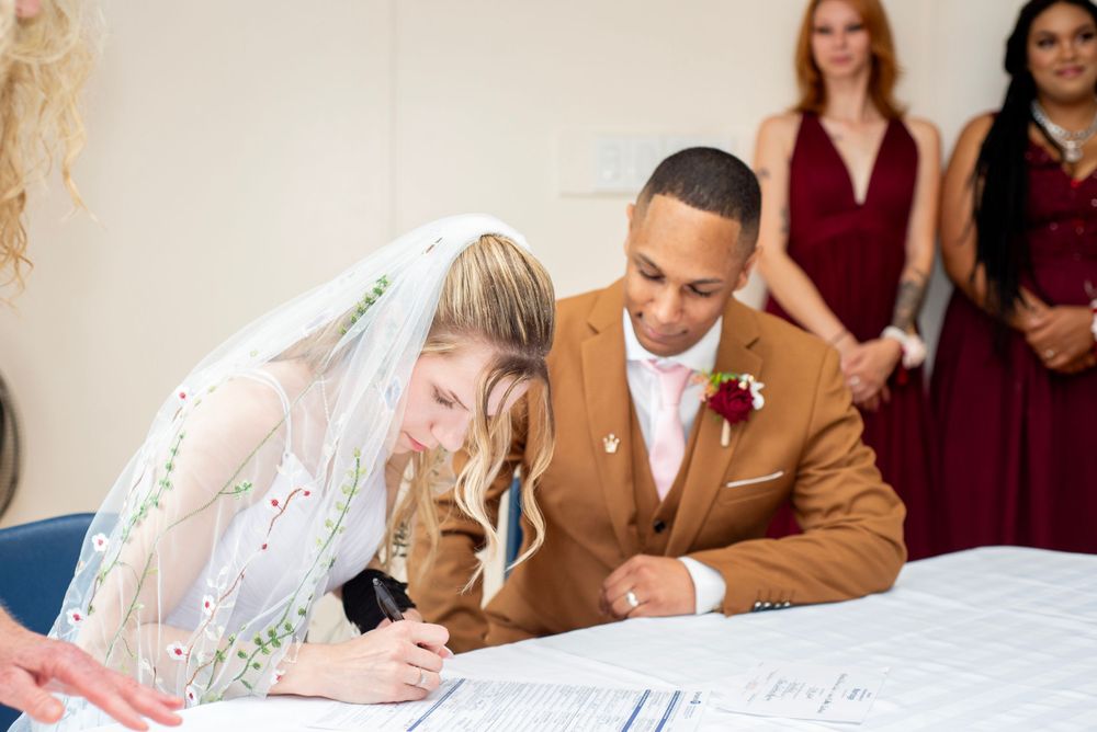 Two people sign marriage documents at a table during a wedding ceremony with bridesmaids in burgundy dresses behind them.