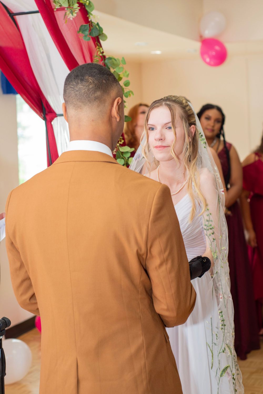 Indoor wedding ceremony with red draped arch and pink balloon decorations.
