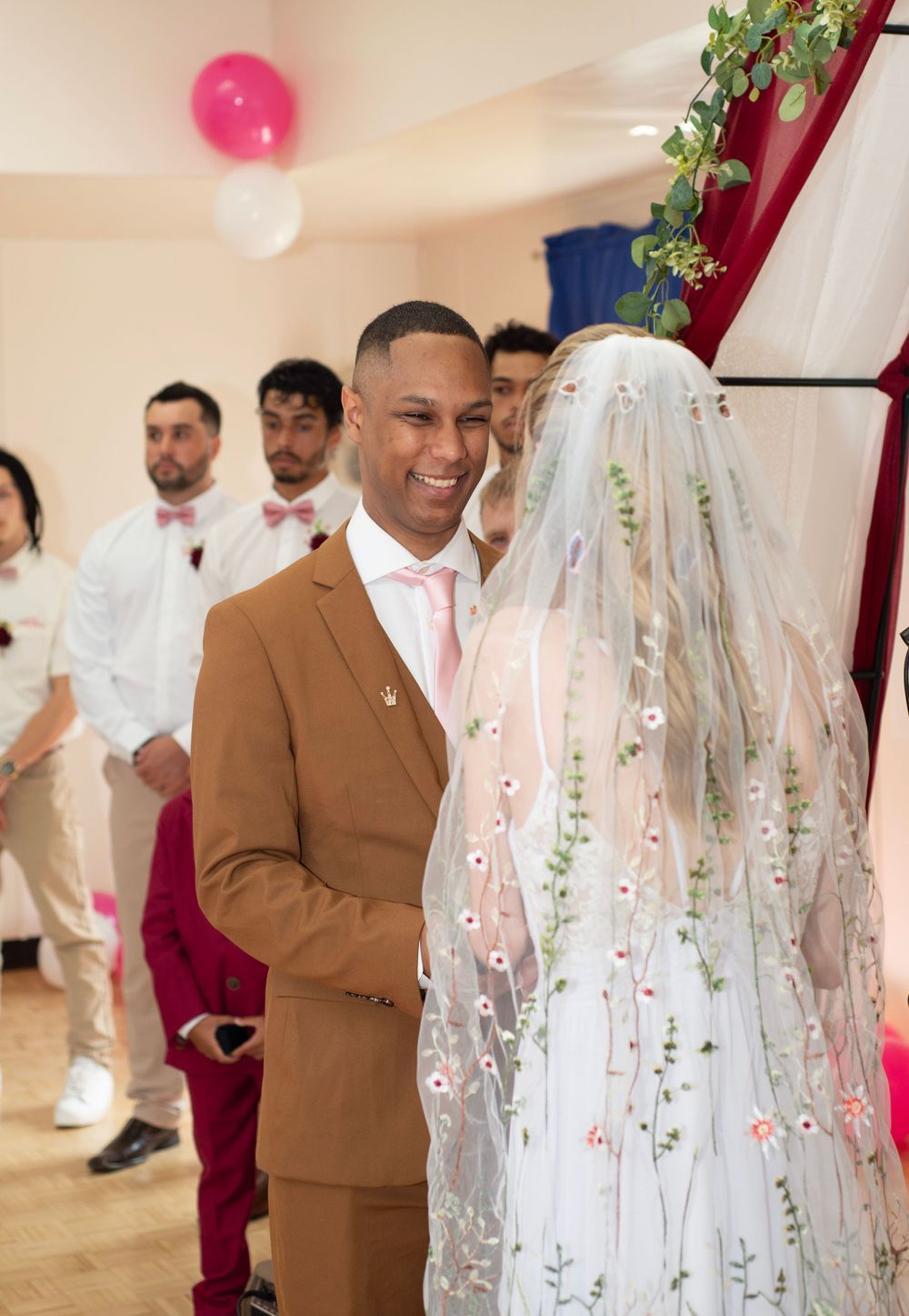 Couple sharing a joyful moment during their indoor wedding ceremony with bridesmaids and groomsmen in the background.