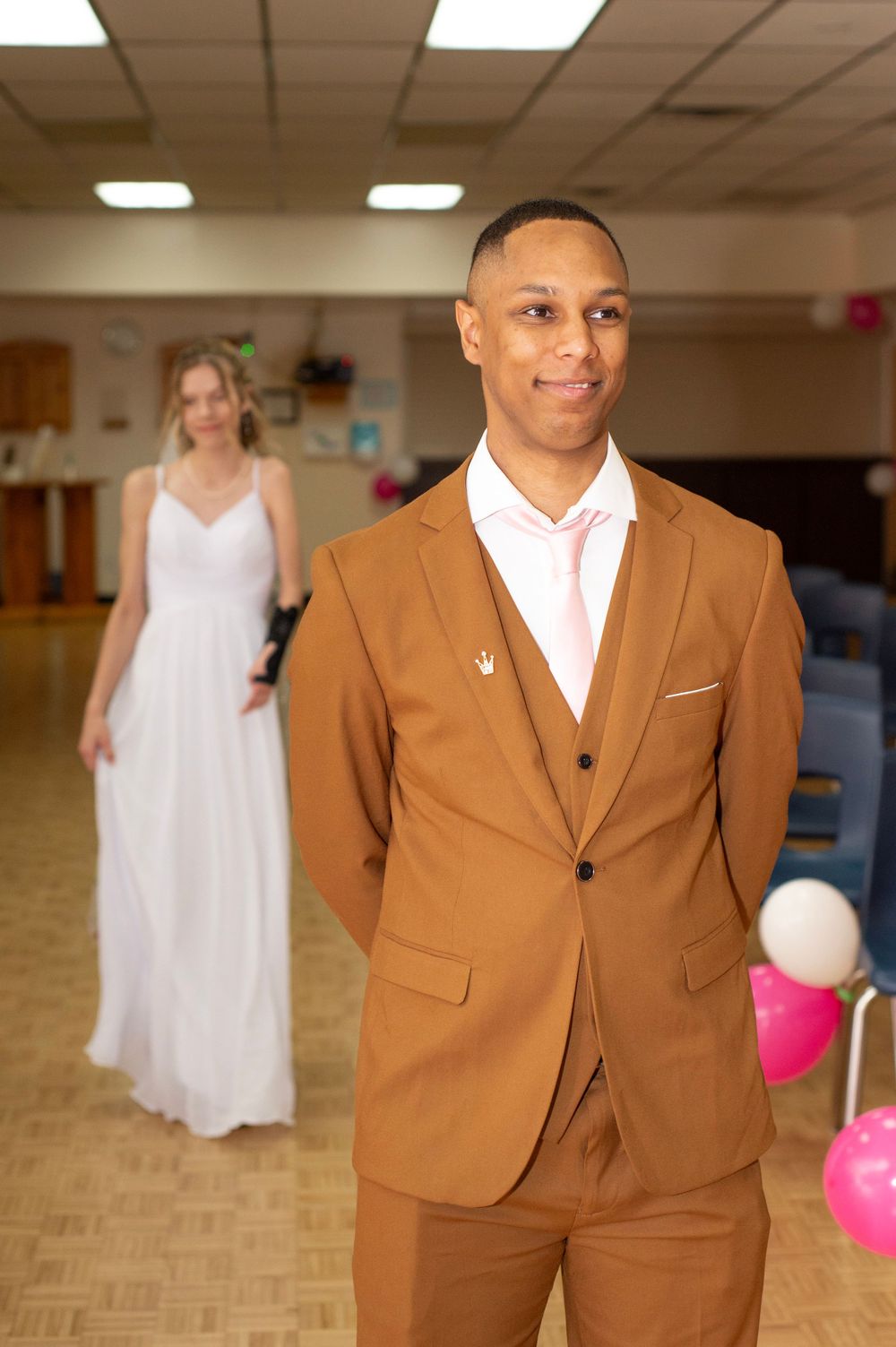 A man in a stylish brown suit poses confidently at an event with decorative balloons and a woman in white behind him.