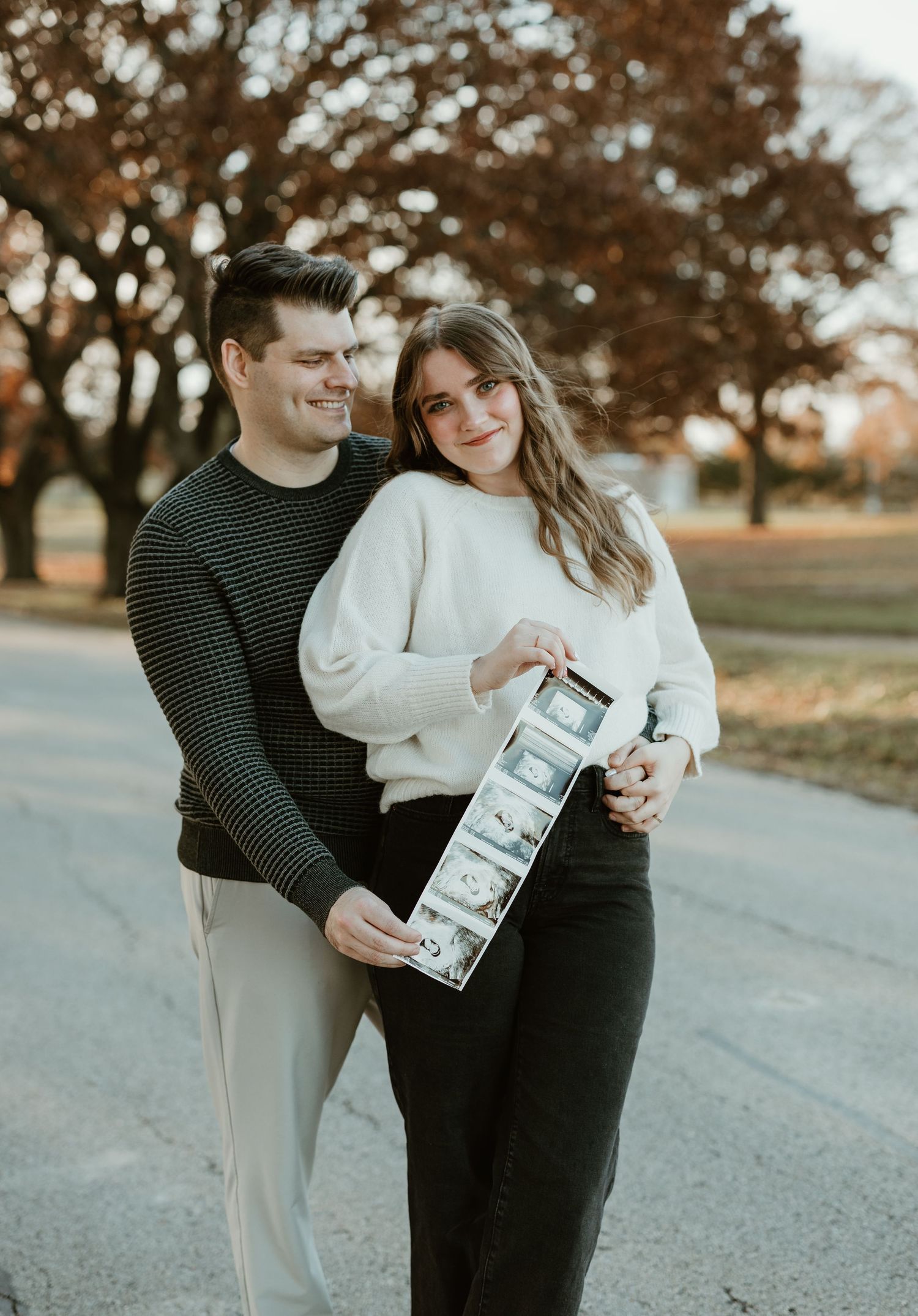 Couple in coordinated sweaters holding ultrasound photos on a pathway.
