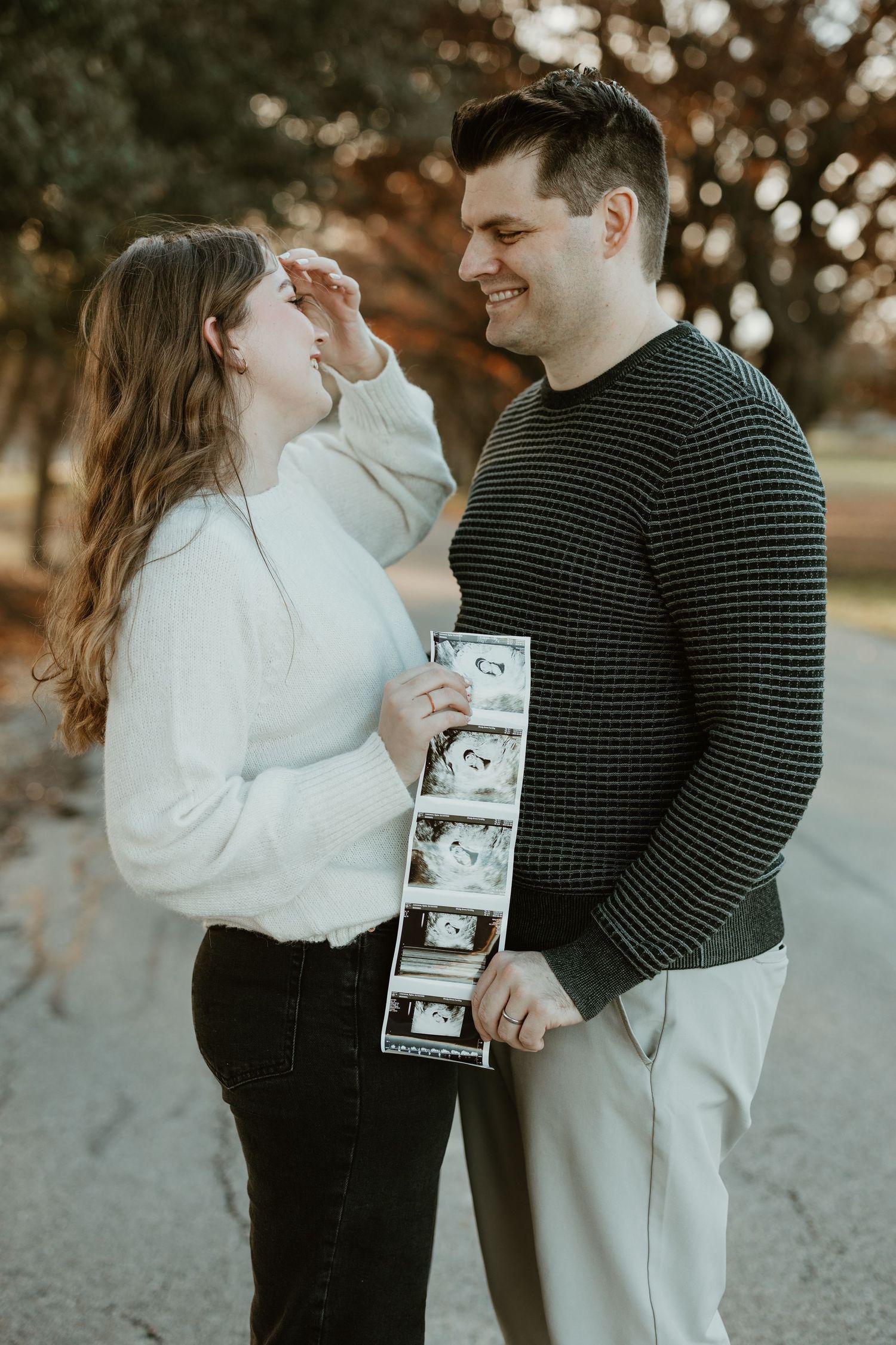 Couple in matching sweaters sharing an emotional moment while holding ultrasound photos outdoors.