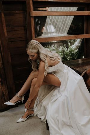 A bride in a white wedding gown adjusts her formal shoes while sitting on rustic wooden steps.