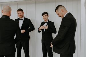 Groomsmen in black tuxedos share casual laughs while getting ready for a wedding ceremony.