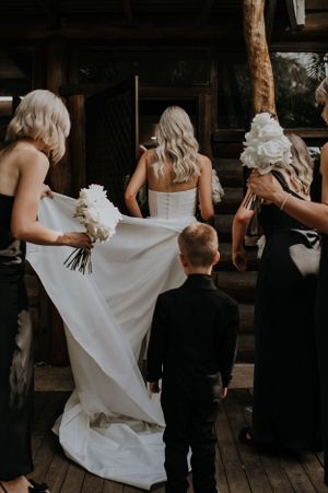 Wedding party members and young attendant gather on wooden deck helping with bridal gown.