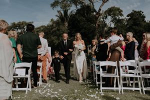 Wedding ceremony guests seated in white chairs on outdoor lawn with flower petals scattered along aisle.
