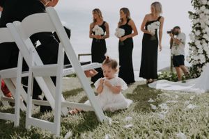 Young flower girl in white dress sits among chairs during outdoor wedding ceremony.