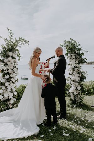 Wedding ceremony under floral arch with white roses and greenery by waterfront.